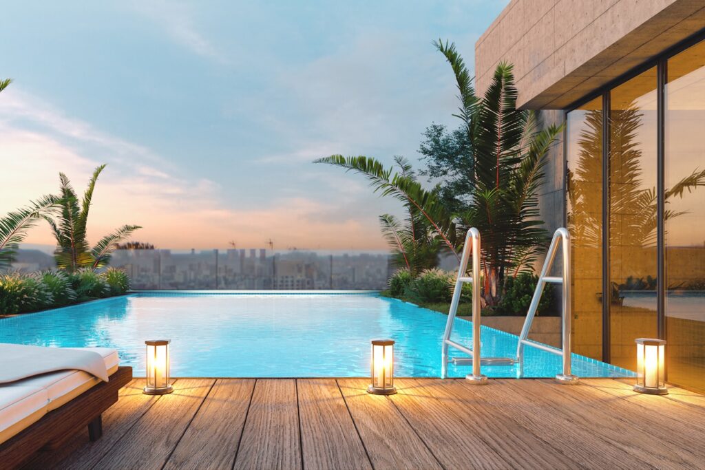 A luxurious rooftop infinity pool at sunrise or sunset. The sleek wooden deck is adorned with modern lounge chairs and warm, ambient lights, creating a serene and inviting space with a scenic view of the city. (This is a computer-generated rendering).